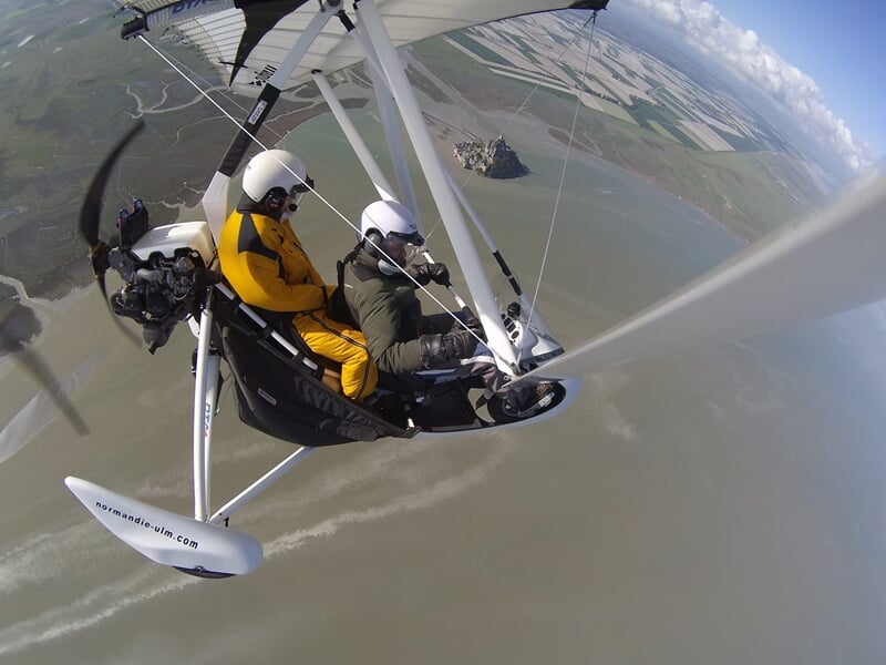 Deux personnes en ULM biplace volant au-dessus d'une zone côtière avec Mont Saint-Michel en arrière-plan.