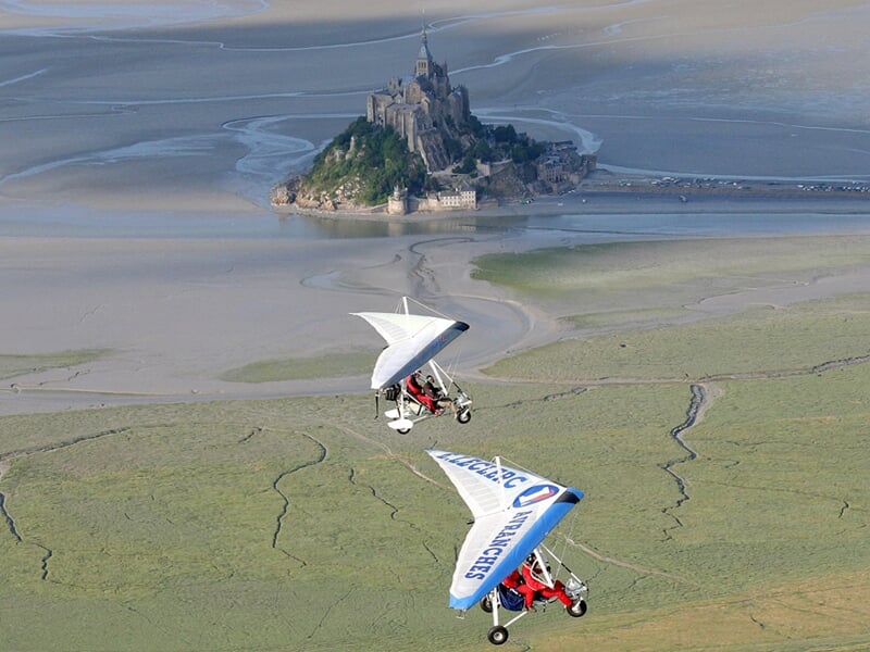 Deux ULM en vol côte à côte au-dessus des terres humides et du Mont Saint-Michel visible au loin.