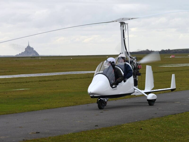 Gyrocoptère au sol sur piste d'atterrissage avec Mont Saint-Michel en arrière-plan sous ciel nuageux.