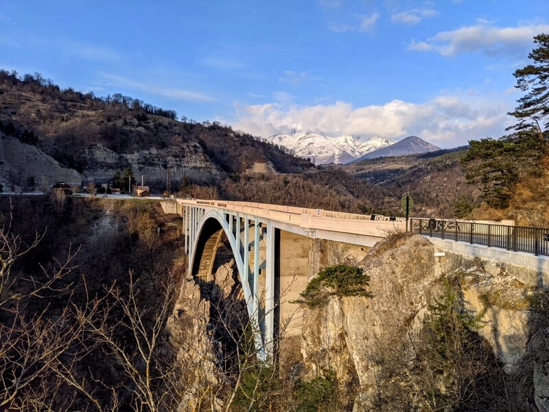 Pont majestueux au-dessus d'une gorge, entouré de montagnes et d'arbres, avec des sommets enneigés en arrière-plan. Lieu prisé pour les sports extrêmes et le tourisme