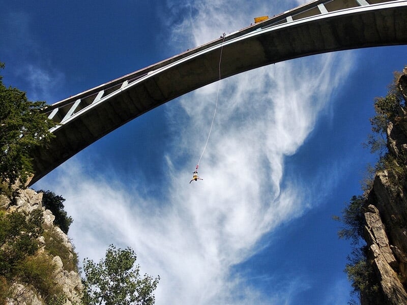 Vue spectaculaire d'un saut à l'élastique sous un pont, une personne suspendue en plein air, ciel bleu et nuages blancs en arrière-plan. Sensations fortes garanties