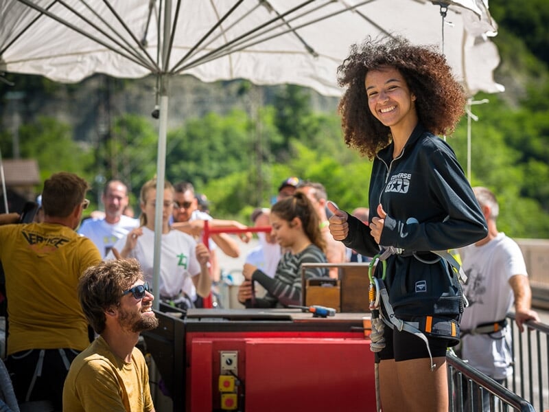 Sourire enthousiaste d'une participante prête pour un saut à l'élastique, portant un harnais de sécurité, entourée de spectateurs et de membres de l'équipe. Ambiance conviviale et sécurisée
