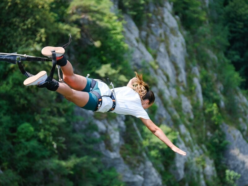 Personne réalisant un saut à l'élastique depuis une hauteur, suspendue par les chevilles. Arrière-plan composé de falaises rocheuses et d'une végétation dense. Tenue sportive : short vert et t-shirt b