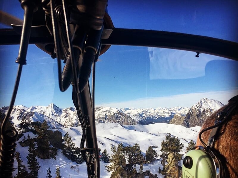 Vue depuis l'intérieur d'un hélicoptère survolant un paysage montagneux enneigé avec un pilote équipé d'un casque.