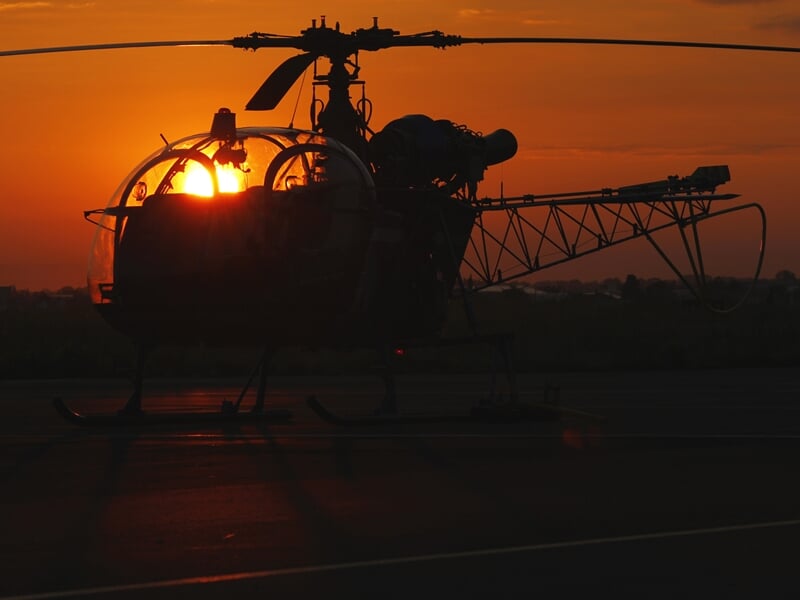 Hélicoptère en contre-jour au coucher du soleil, ses contours et ses pales se détachant dans une lumière orange intense, symbolisant l'aviation romantique ou les vols en soirée