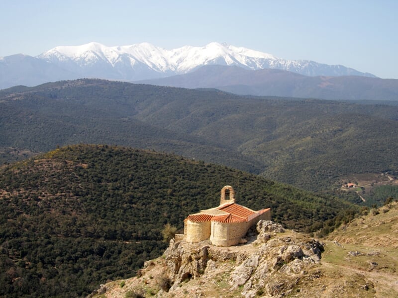 Chapelle isolée sur une colline verdoyante avec en arrière-plan des montagnes enneigées sous un ciel clair.