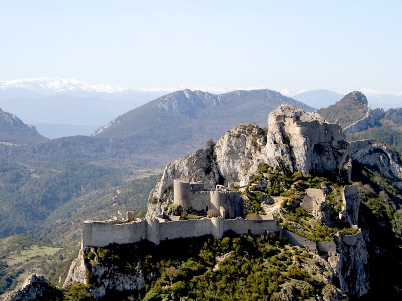 Ruines d'un château fort perché sur une colline rocheuse entourée de végétation et de montagnes au loin.