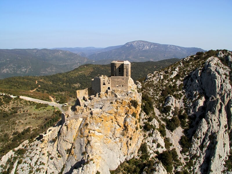 Château perché sur une falaise rocheuse avec une vue panoramique sur des collines verdoyantes et un ciel clair, capturé depuis un hélicoptère