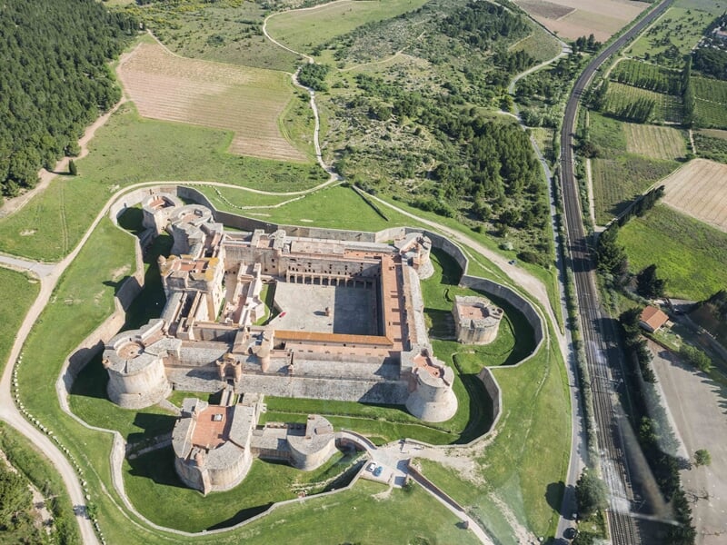 Vue aérienne d'une forteresse entourée de douves et de verdure, avec une route et des champs à proximité.