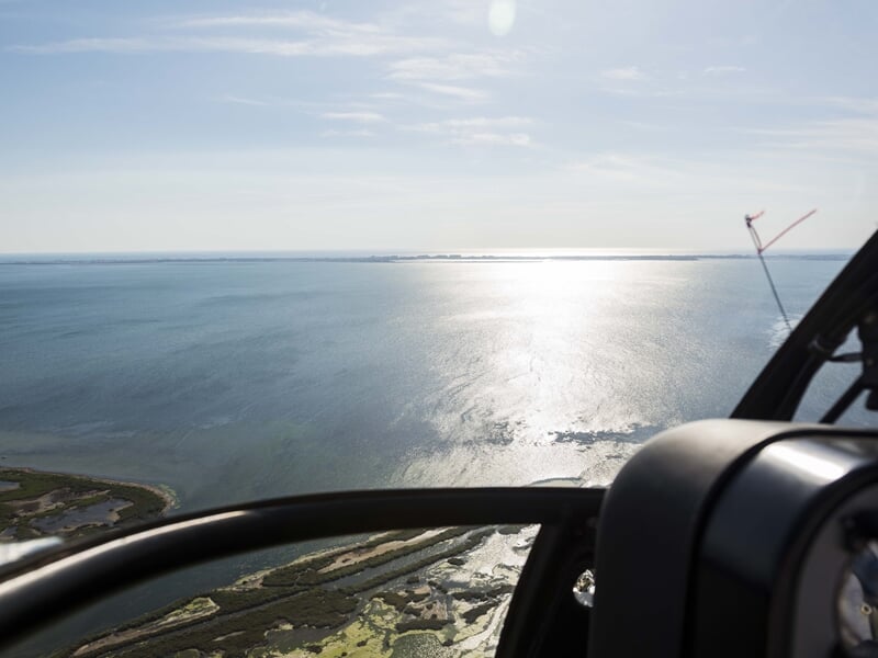 Vue aérienne depuis un hélicoptère sur une étendue d'eau scintillante sous le soleil, bordée par des zones humides et des îles, parfaite pour illustrer des tours en hélicoptère
