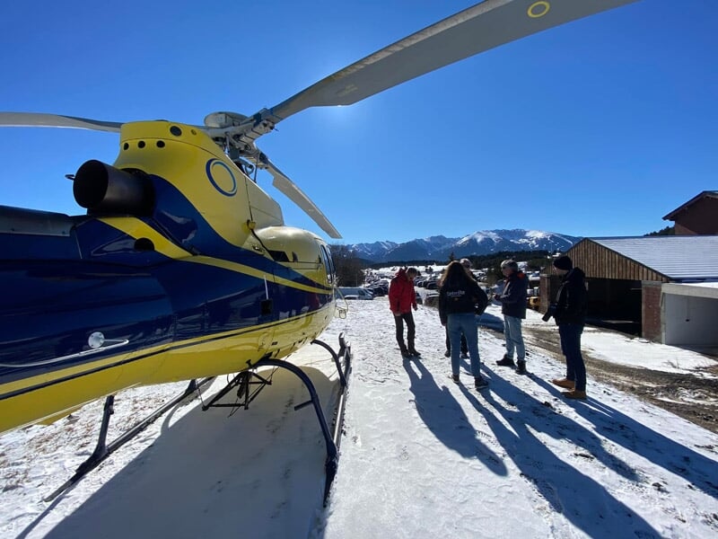 Hélicoptère jaune et bleu posé sur la neige avec plusieurs personnes debout à côté, montagnes en arrière-plan.