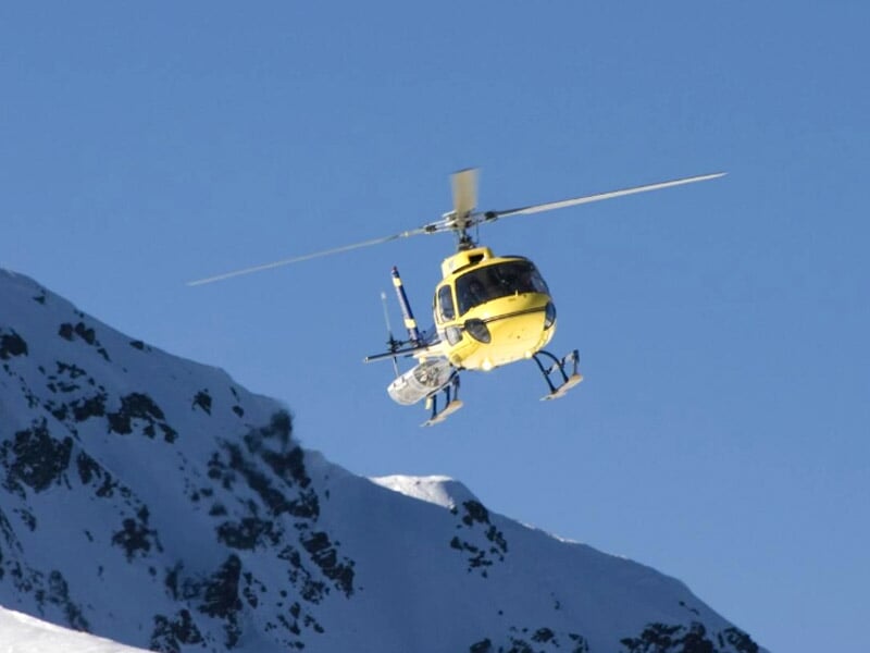 Hélicoptère jaune en vol au-dessus d'une montagne enneigée sous un ciel bleu clair sans nuages.