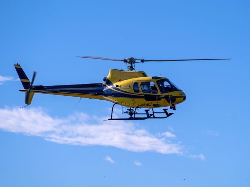 Hélicoptère jaune et bleu en vol dans un ciel dégagé, ses pales en mouvement au-dessus d'un nuage léger, utilisé pour des missions aériennes ou de transport