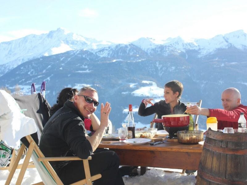 Groupe de personnes partageant un repas en plein air sur une terrasse enneigée avec montagnes en arrière-plan.