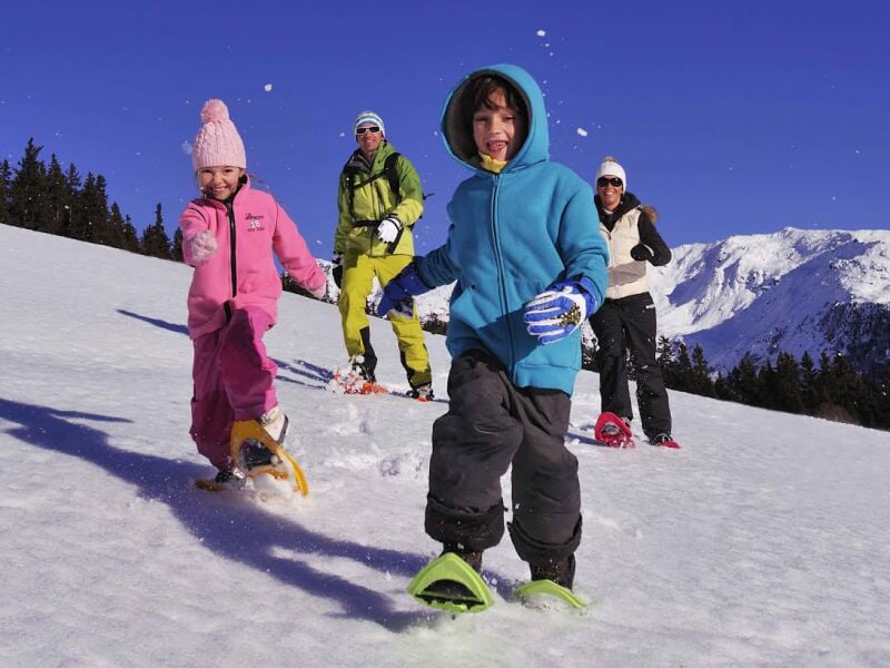 Famille joyeuse marchant dans la neige avec des raquettes, sous un ciel bleu et des montagnes en arrière-plan.