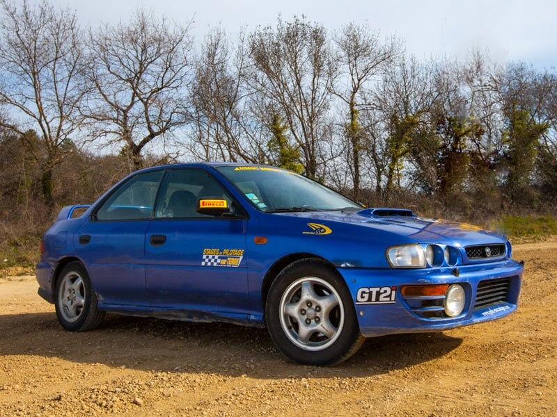 Voiture de rallye bleue stationnée sur une piste de terre avec des arbres en arrière-plan