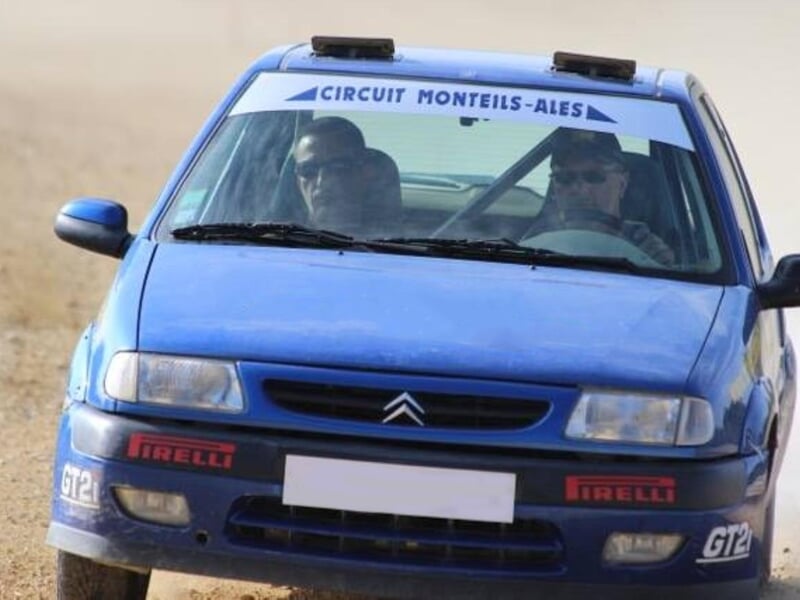 Voiture de rallye bleue avec deux personnes à bord sur une piste de terre, vue de face