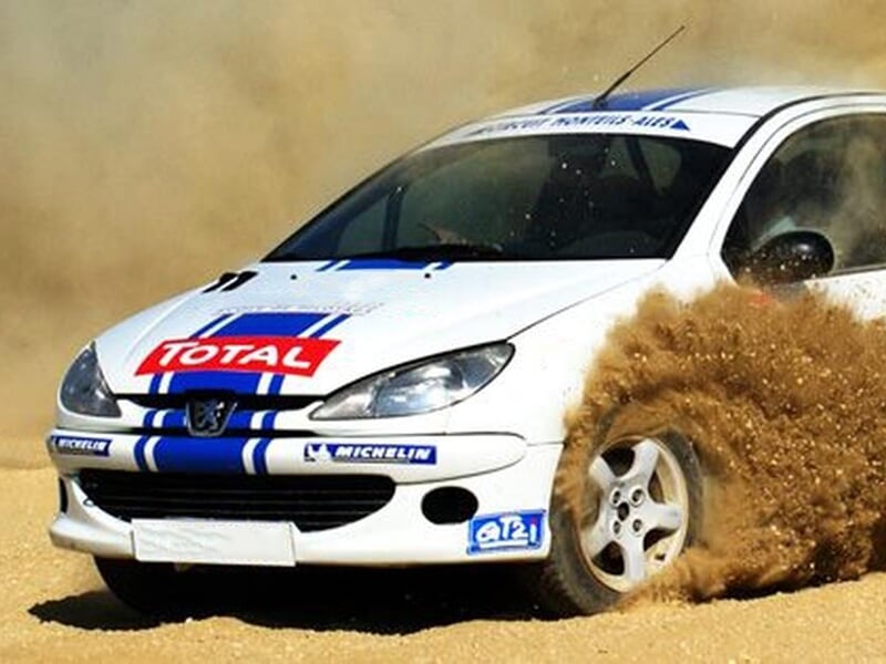 Voiture de rallye blanche avec des bandes bleues dérapant sur une piste de terre, soulevant de la poussière