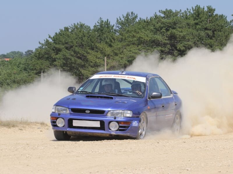 Voiture de rallye bleue dérapant sur une piste de terre, avec deux personnes à bord