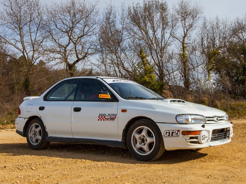 Voiture de rallye bleue sur une piste de terre avec des arbres sans feuilles en arrière-plan