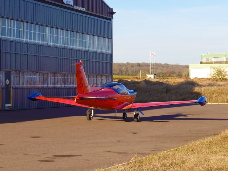 Petit avion rouge et bleu stationné devant un hangar à l'aéroport de Strasbourg par temps clair.