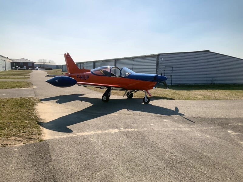 Avion rouge et bleu sur une piste d'aéroport avec hangars en arrière-plan sous un ciel dégagé.