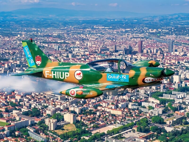Avion de voltige vert avec motifs colorés volant au-dessus d'une grande ville sous un ciel bleu.