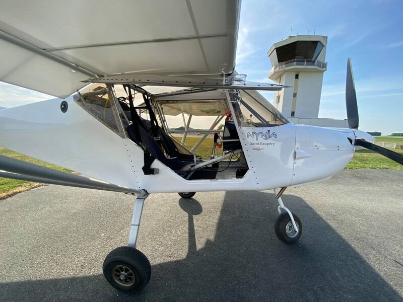 Avion ULM blanc stationné au sol avec porte ouverte et tour de contrôle en arrière-plan.