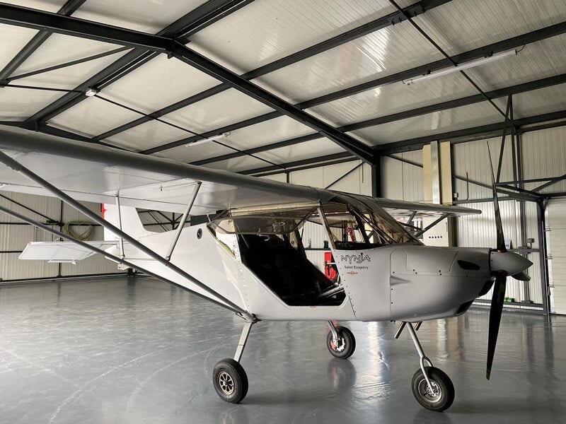 Avion ULM blanc garé dans un hangar avec lumière naturelle et sol en béton.