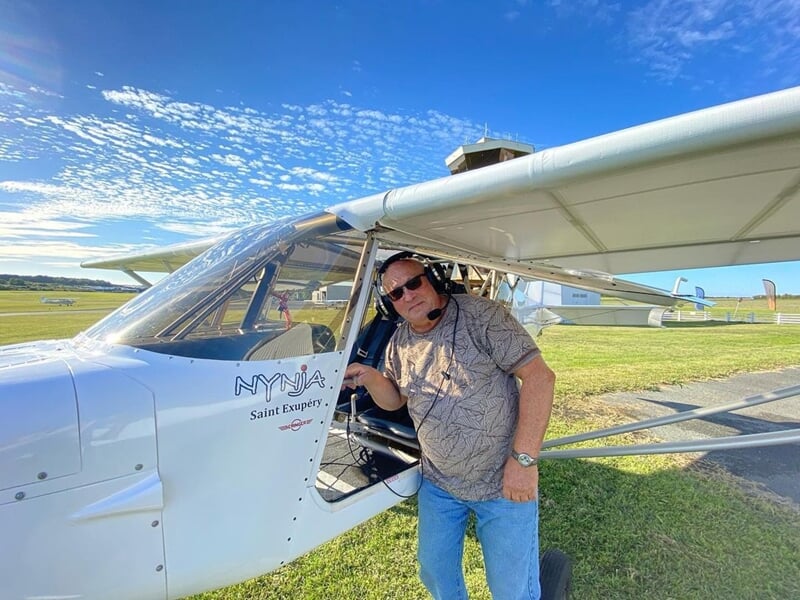 Homme debout à côté d'un avion ULM blanc sur une piste d'atterrissage ensoleillée.