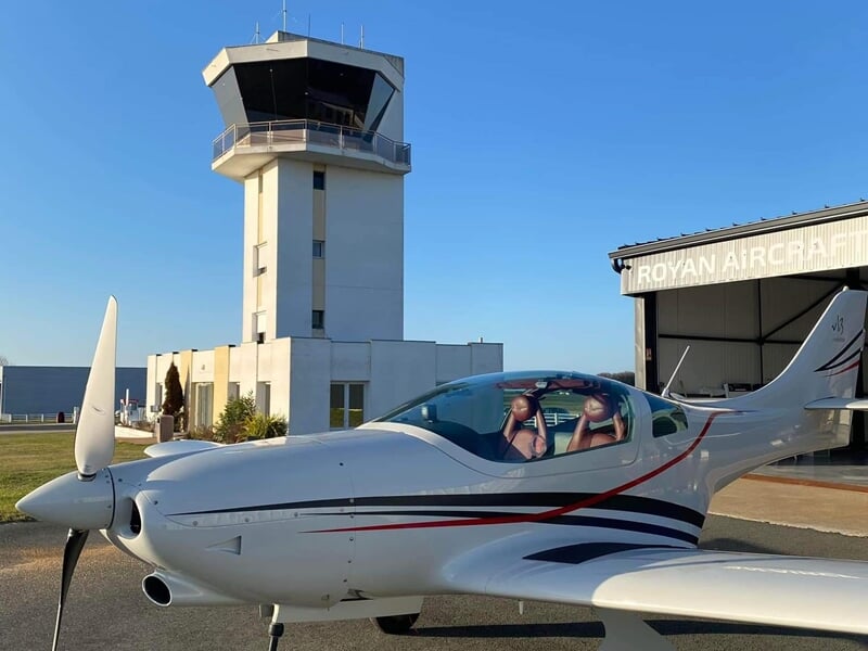 Petit avion de tourisme avec deux personnes à bord devant un hangar et une tour de contrôle.