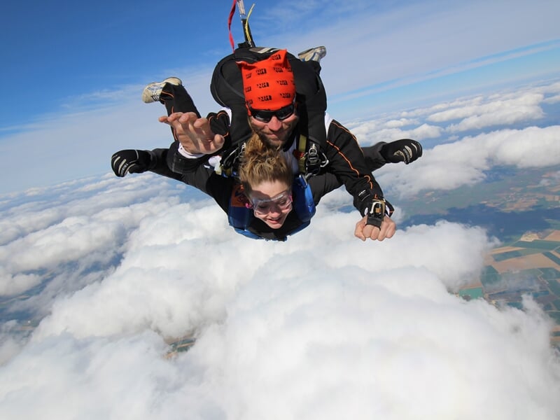 Deux personnes en chute libre lors d'un saut en parachute au-dessus des nuages et du paysage terrestre.