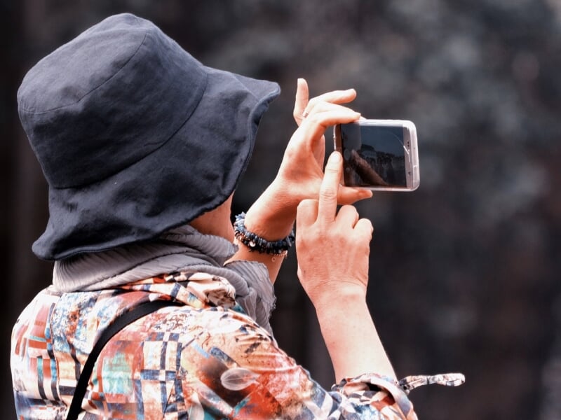 Une personne en chapeau et vêtements colorés prenant une photo avec un smartphone. L'image capture un moment de photographie en extérieur.