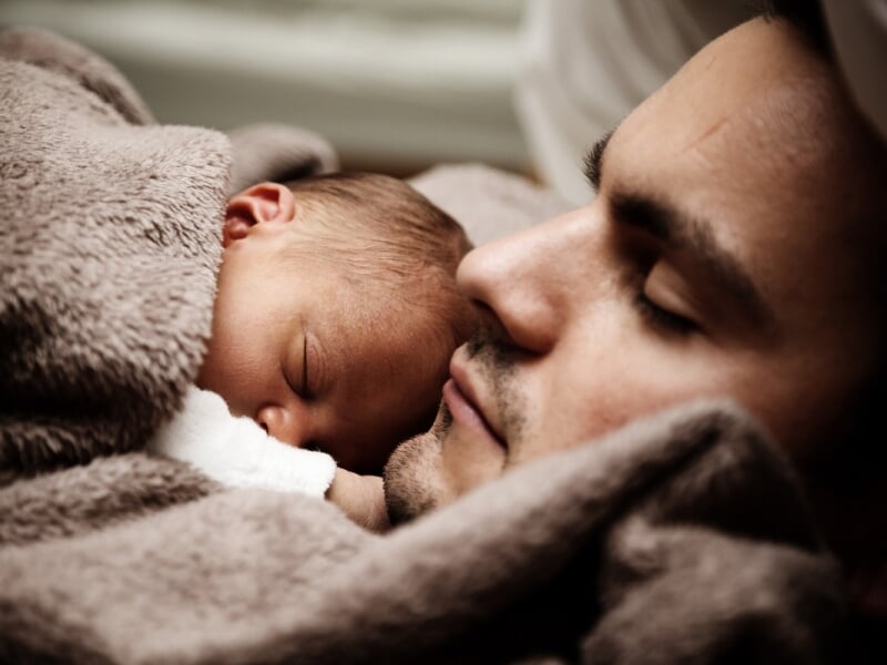 Un homme endormi avec un bébé blotti contre lui, enveloppés dans une couverture douce et beige. L'image évoque tendresse et lien parental.