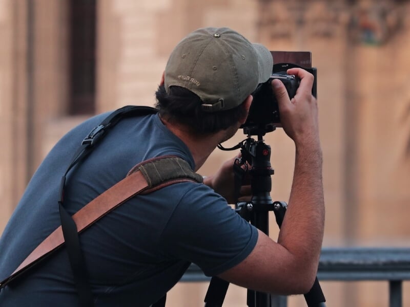 Un homme photographiant avec un appareil photo sur trépied, portant une casquette et un t-shirt bleu. L'arrière-plan flou met en évidence le photographe en action.