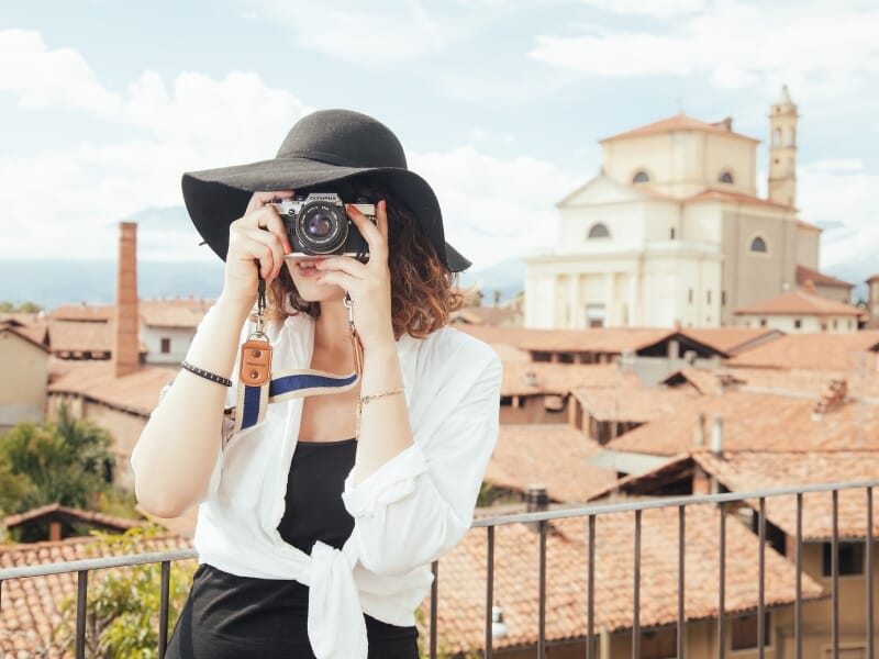 Femme portant un chapeau noir et une chemise blanche, photographiant avec un appareil photo vintage sur une terrasse, avec des toits de tuiles rouges et une église en arrière-plan. La scène évoque un 