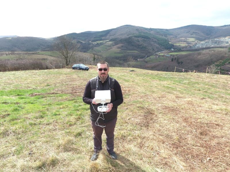 Homme tenant une télécommande de drone, debout dans un champ herbeux avec des collines verdoyantes et un véhicule stationné à l'arrière-plan