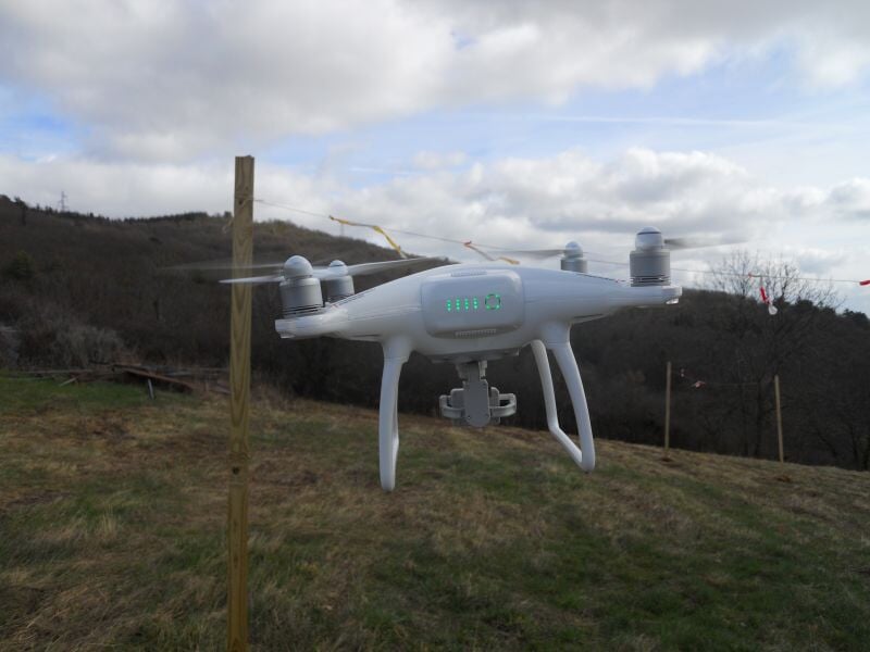 Drone blanc en vol stationnaire en plein air, affichant des indicateurs LED verts sur son châssis, au-dessus d'un champ herbeux avec des collines en arrière-plan