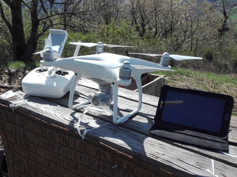 Drone blanc avec télécommande et tablette posés sur une surface en bois, prêt à décoller dans un environnement naturel avec arbres en arrière-plan