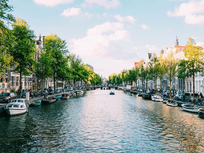 Vue d'un canal urbain avec des bateaux amarrés, des arbres verts et des bâtiments sous un ciel bleu clair.