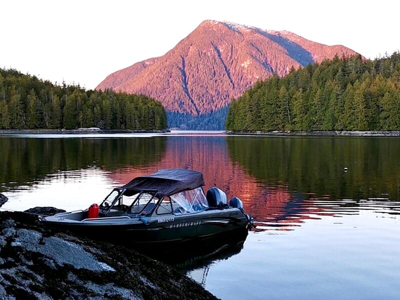 Petit bateau noir amarré au bord d'un lac calme avec une montagne rose en arrière-plan et une forêt dense.