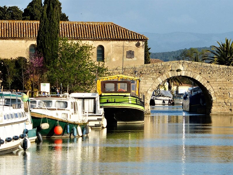 Canal avec plusieurs bateaux amarrés, un pont en pierre et des bâtiments anciens sous un ciel clair.