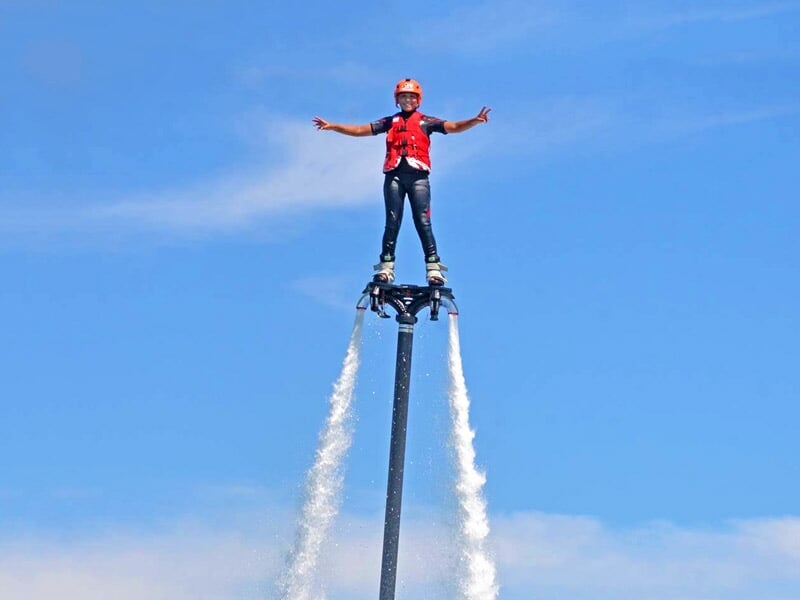 Homme en flyboard avec casque et gilet rouge, en équilibre sur l'eau sous un ciel bleu clair.