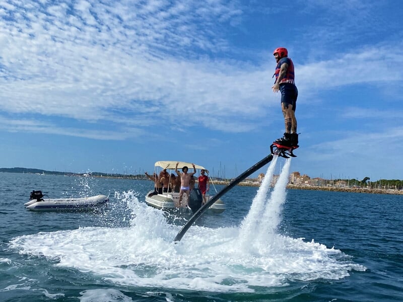 Homme en flyboard près d'un bateau avec plusieurs personnes, sous un ciel partiellement nuageux.
