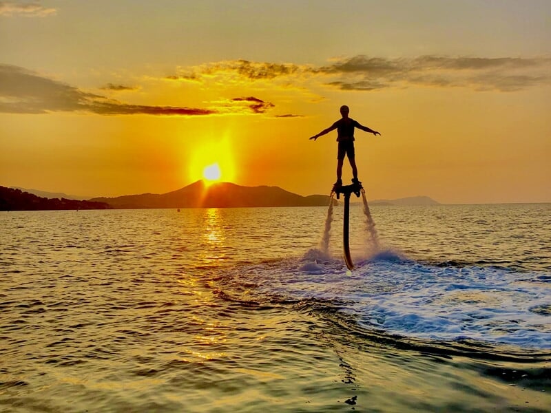 Flyboarder silhouetté au coucher du soleil, sur la mer calme avec des montagnes à l'horizon.