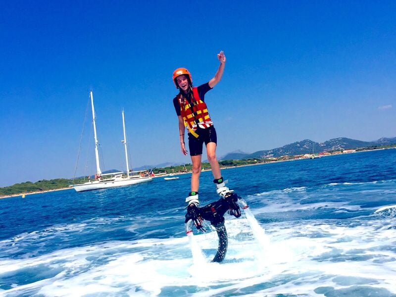 Personne en flyboard sur l'eau, portant un casque et un gilet de sauvetage, avec un voilier en arrière-plan.