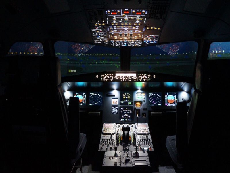 Simulateur de pilotage d'avion avec écrans et commandes illuminés dans un cockpit sombre.