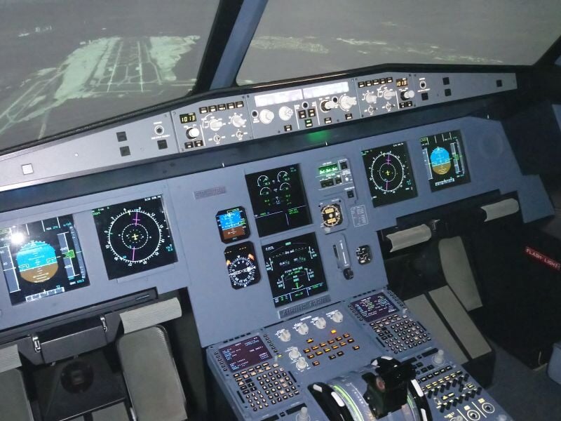 Cockpit d'un simulateur de pilotage d'avion avec plusieurs écrans et commandes numériques en vue intérieure.