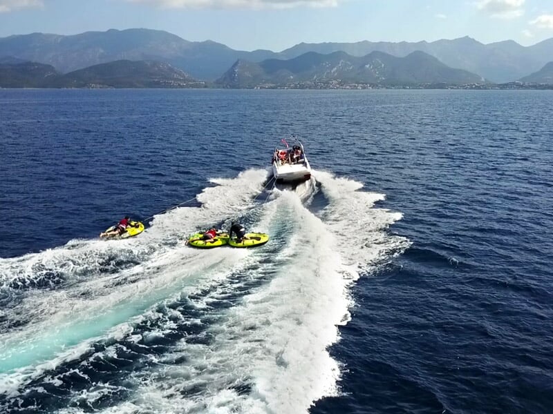 Bateau tirant trois bouées tractées avec des personnes s'amusant sur l'eau en mer calme et montagnes en arrière-plan.