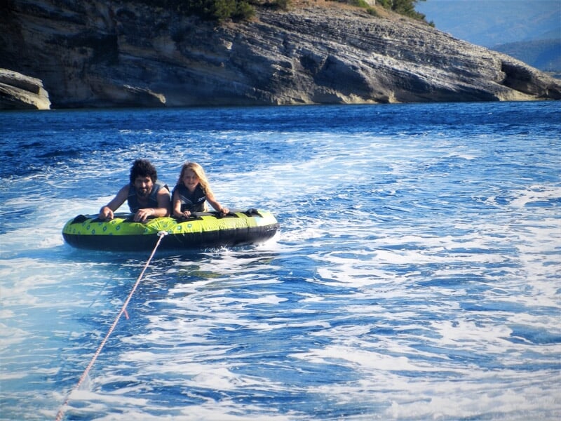 Deux personnes sur une bouée tractée sur l'eau bleue, entourées de rochers et de vagues légères sous un ciel clair.